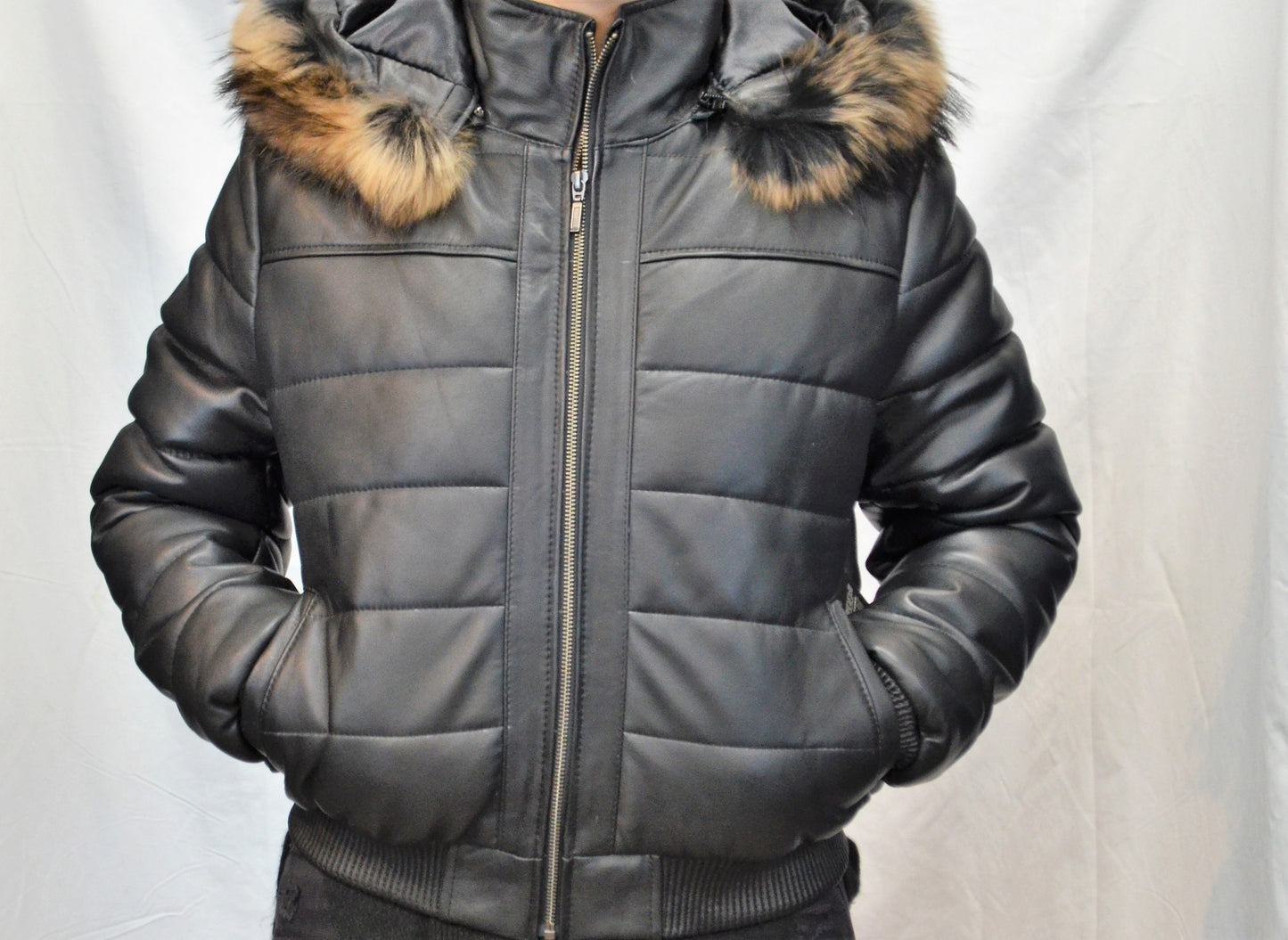 Front view of leather jacket on model with light puffer design and real fur hood, zipped up and hands in pocket. 