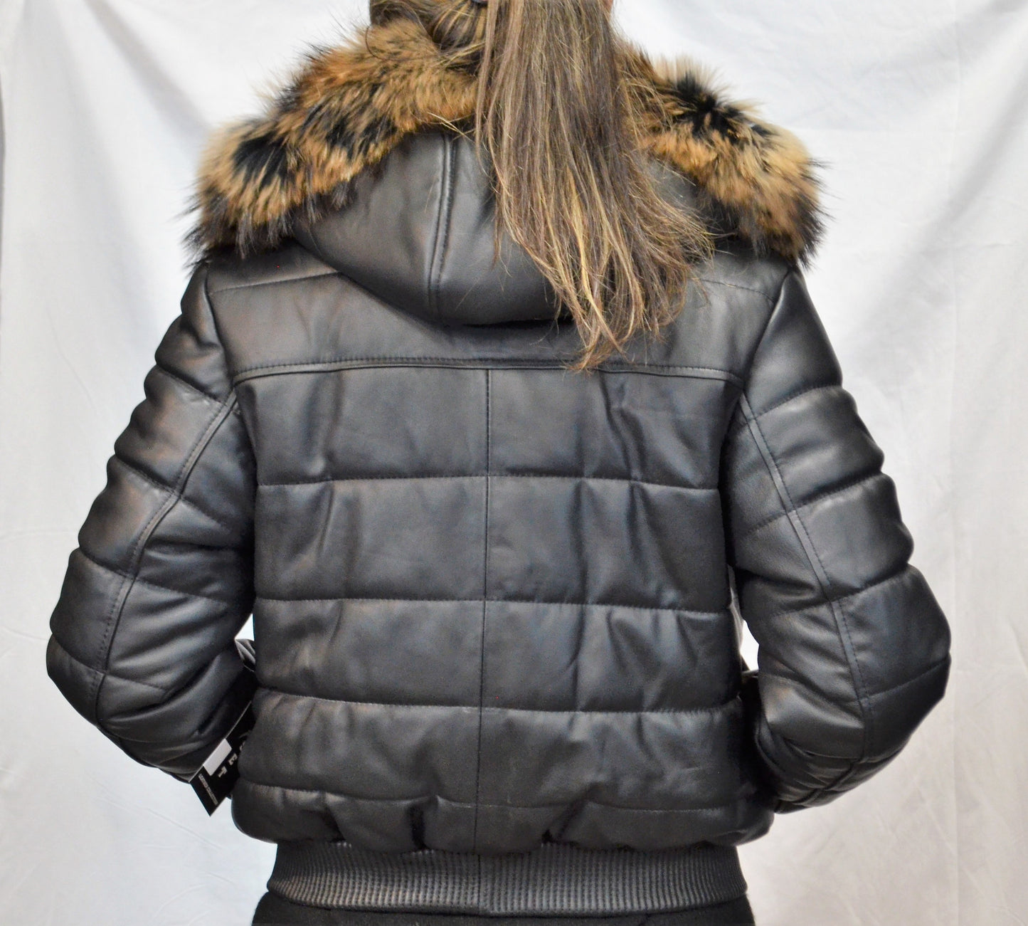 Back view of leather jacket on model with light puffer design and real fur hood, hands in pocket. 